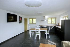 A modern kitchen with a dining area, featuring a large flat-screen TV, a wooden dining table, and white chairs.
