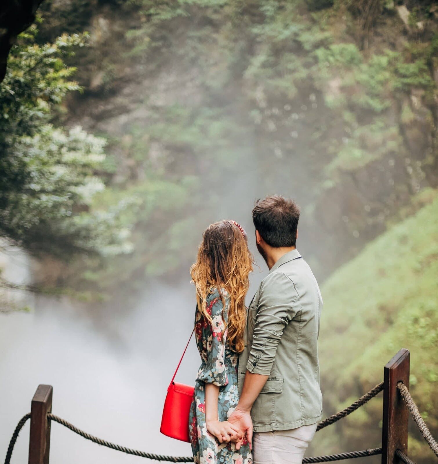 A couple standing on a rope bridge with a forested river in the background.