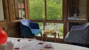 A room with a bed, a table with a fruit platter, a chair, and a window with a view of greenery.