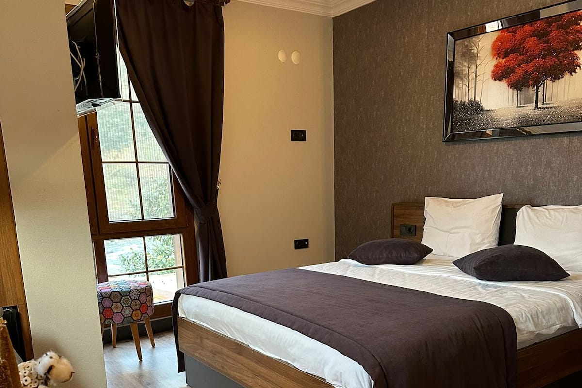 A bedroom with a wooden bed, a window with curtains, a painting on the wall, and a small table with decorative items.