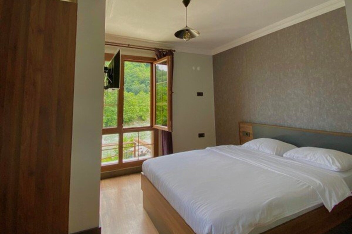 A bedroom with a large bed, wooden flooring, a ceiling fan, and a window with a view of greenery.