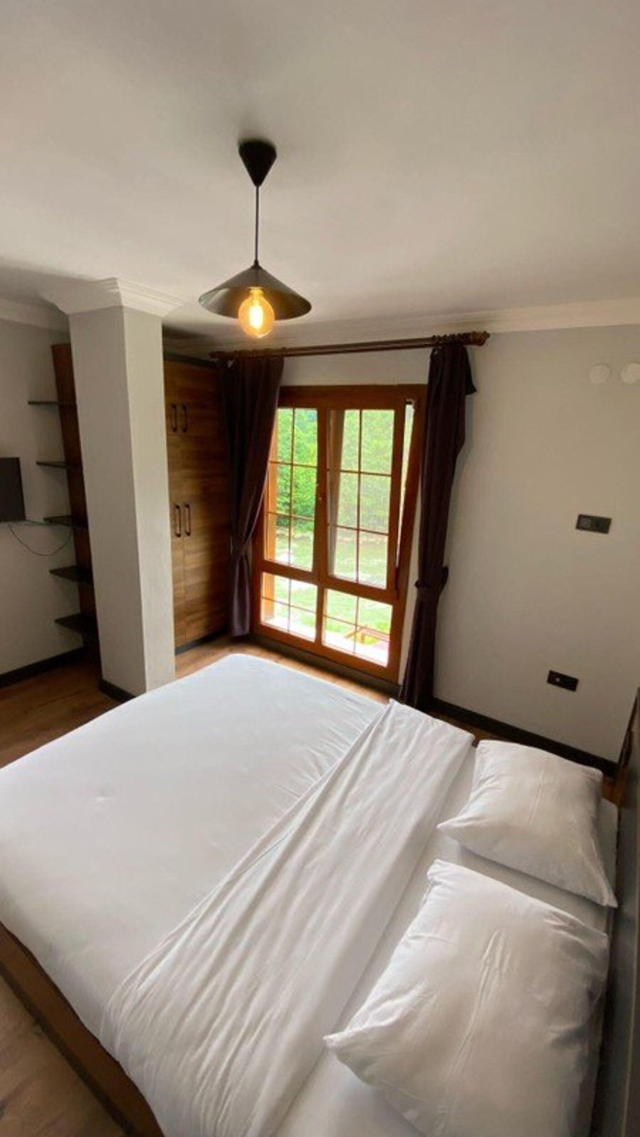 A bedroom with a large bed, a ceiling light, a window with curtains, and a wooden wardrobe.
