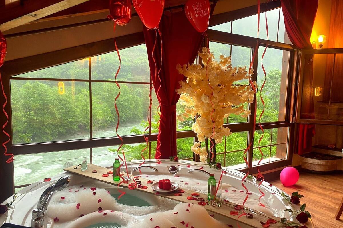 A room with a large window, a bathtub with rose petals, and a vase with white flowers.