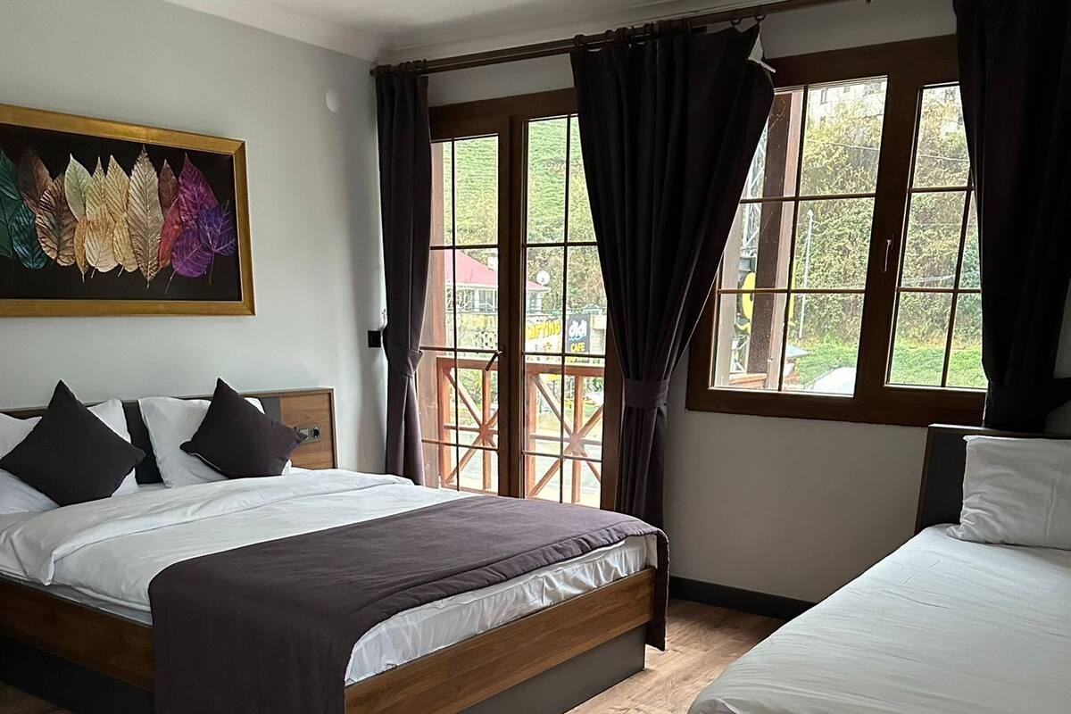 A hotel room with two beds, a wooden floor, a dining area with a chair, and large windows with curtains.