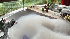A bathroom with a bathtub, a window with a view of a forest and river, and a table with a glass of water, a rose, and some fruit.