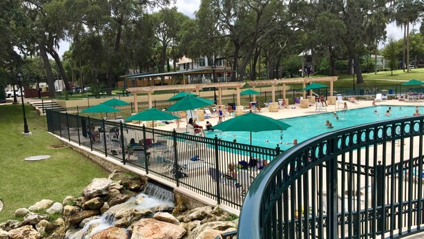 A swimming pool with a waterfall feature, surrounded by a fence and shaded by umbrellas. - Historic Green Cove Springs Home - Walk to Coast! (Green Cove Springs)