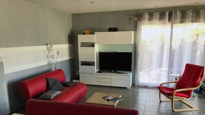 A modern living room with a red sofa, a TV on a white cabinet, a red chair, and a view of the outdoors through a window.