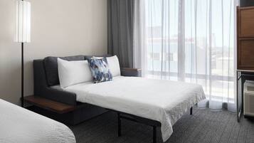 Quarto de hotel com cama, sofá, luminária, janela com cortinas e vista da cidade.
