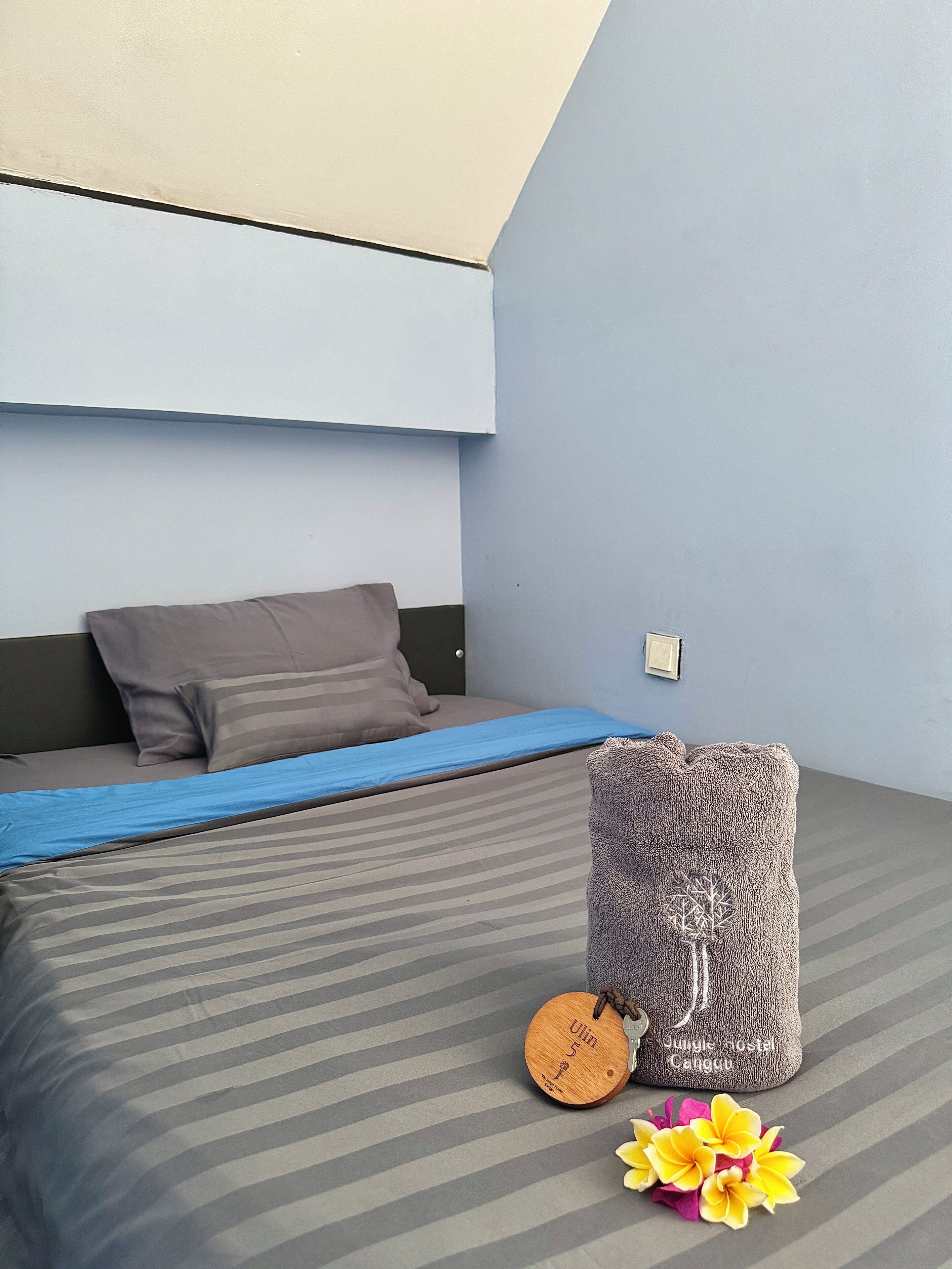 A bedroom with a bed, pillows, a blue blanket, a wooden tag, a plush cushion, and a small wooden stool.