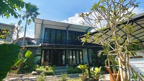 A modern building with a covered patio area, surrounded by various plants and a stone pathway. - The Jungle Hostel Canggu (Pererenan)