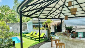 A covered outdoor area with lounge chairs, a pool, and a bar with stools. - The Jungle Hostel Canggu (Pererenan)