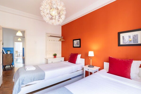 Habitación de hotel con dos camas, una mesita de noche blanca, un aparador blanco y una pared roja, además de un cuadro en la pared.