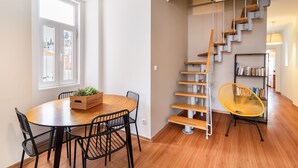 Un comedor con una mesa redonda de madera y sillas negras, una escalera de madera con barandilla de metal, y una estantería llena de libros.