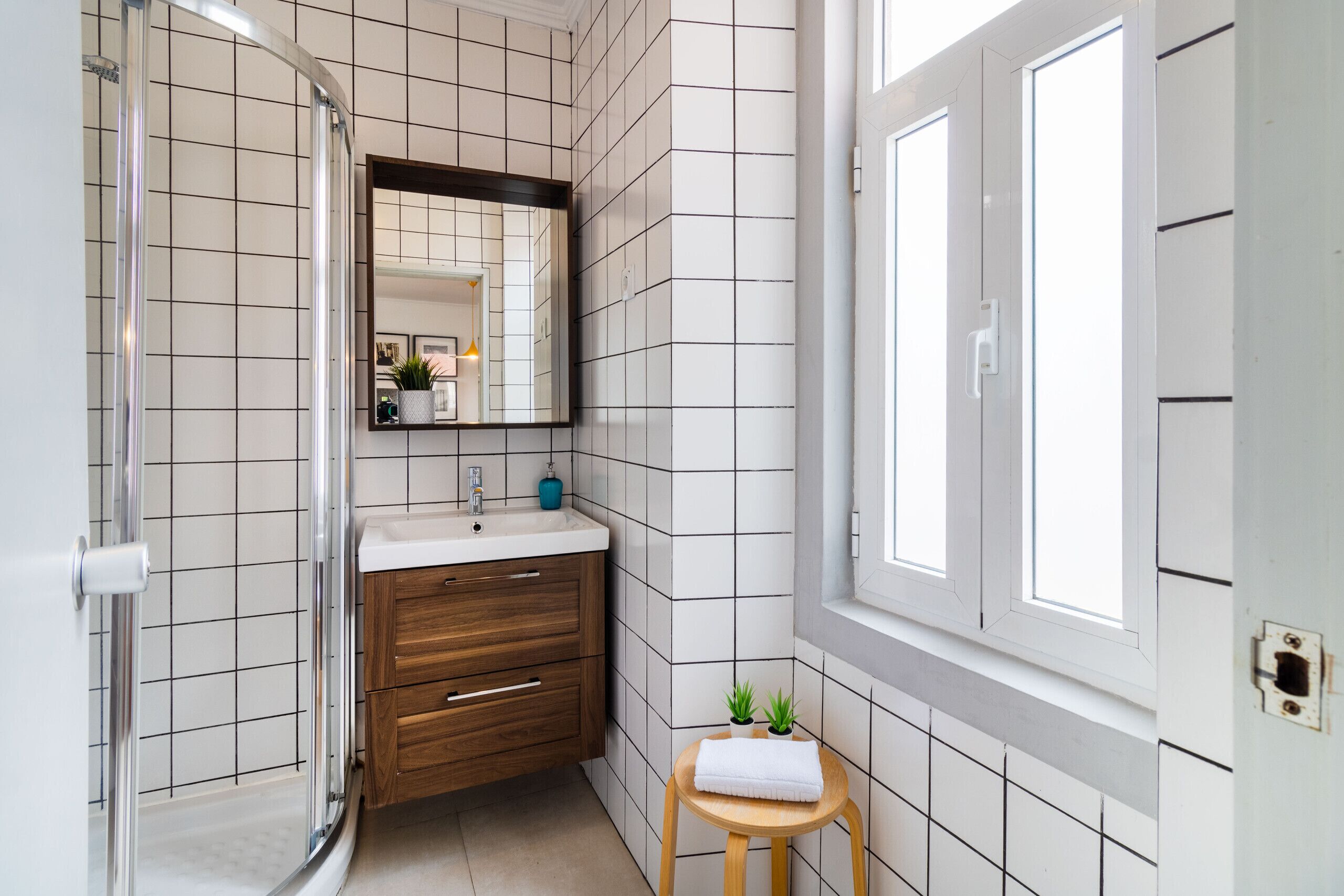 A bathroom with a glass shower enclosure, a wooden vanity with a sink, and a small stool with a towel.
