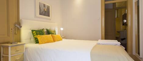 A neatly made bed with white and yellow bedding, a bedside lamp, and a framed picture on the wall.