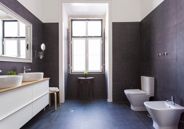A modern bathroom with dark tiled walls, a large mirror, a sink, a toilet, and a bidet.