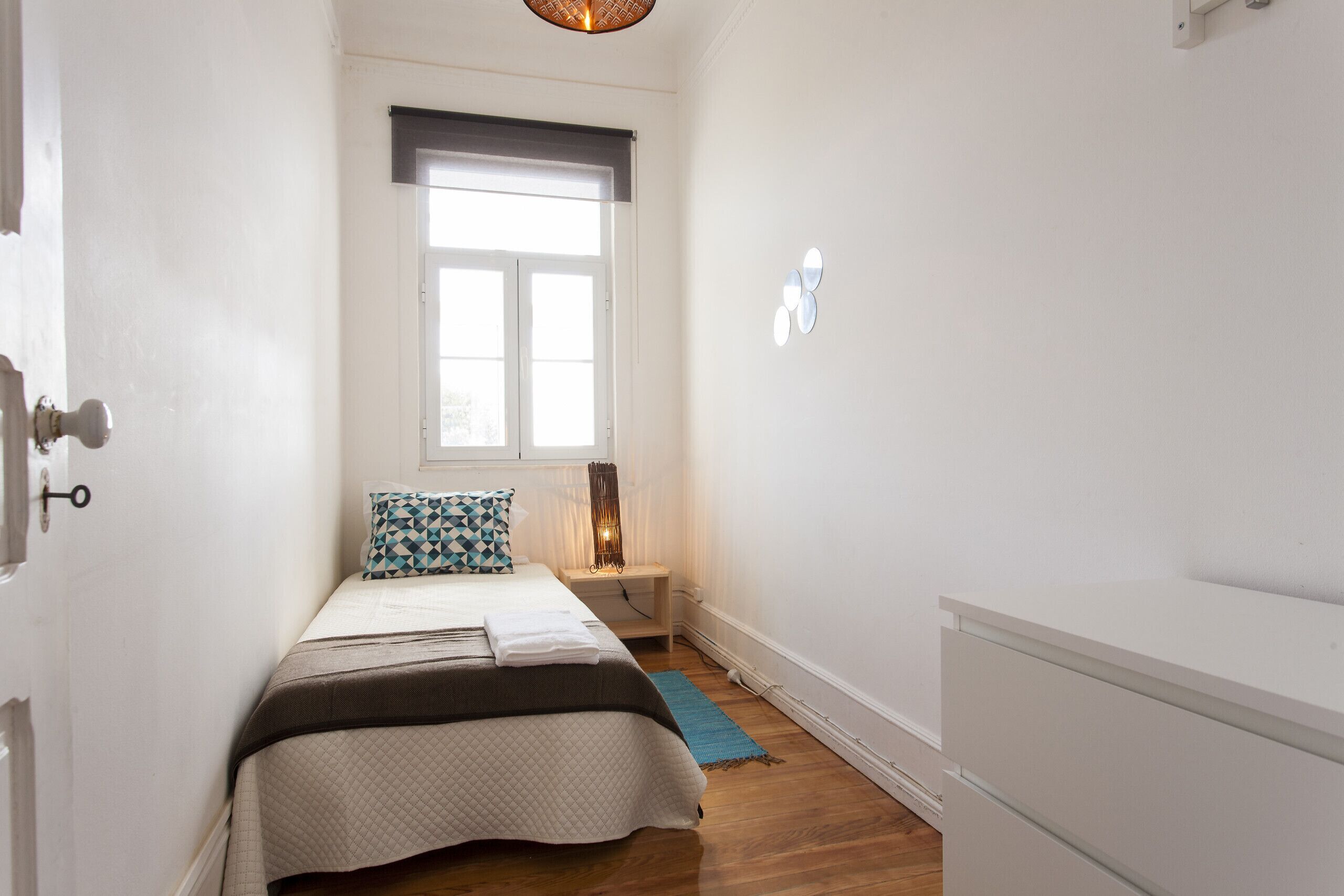 Ein Schlafzimmer mit einem Bett, einem Fenster mit Jalousien, einem Nachttisch mit Lampe und einer Kommode.