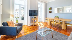 Ein Wohnzimmer mit einer Couch, einem Glastisch, einem Fernseher, einem Esstisch mit Stühlen und einem Bücherregal.