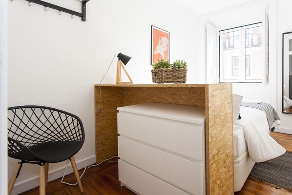 A compact room with a wooden desk, a black chair, a white dresser, a wicker basket with plants, and a framed picture on the wall.