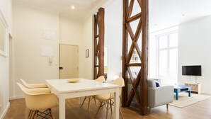 Una sala moderna con un panel de madera, una mesa de comedor blanca con sillas, un sofá gris y un televisor de pantalla plana.