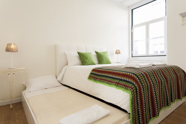 A neatly made bed with white linens and green pillows, a patterned throw blanket, and a bedside table with a lamp.
