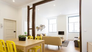 A bright living room with a wooden dining table, yellow chairs, a sofa, and a TV.