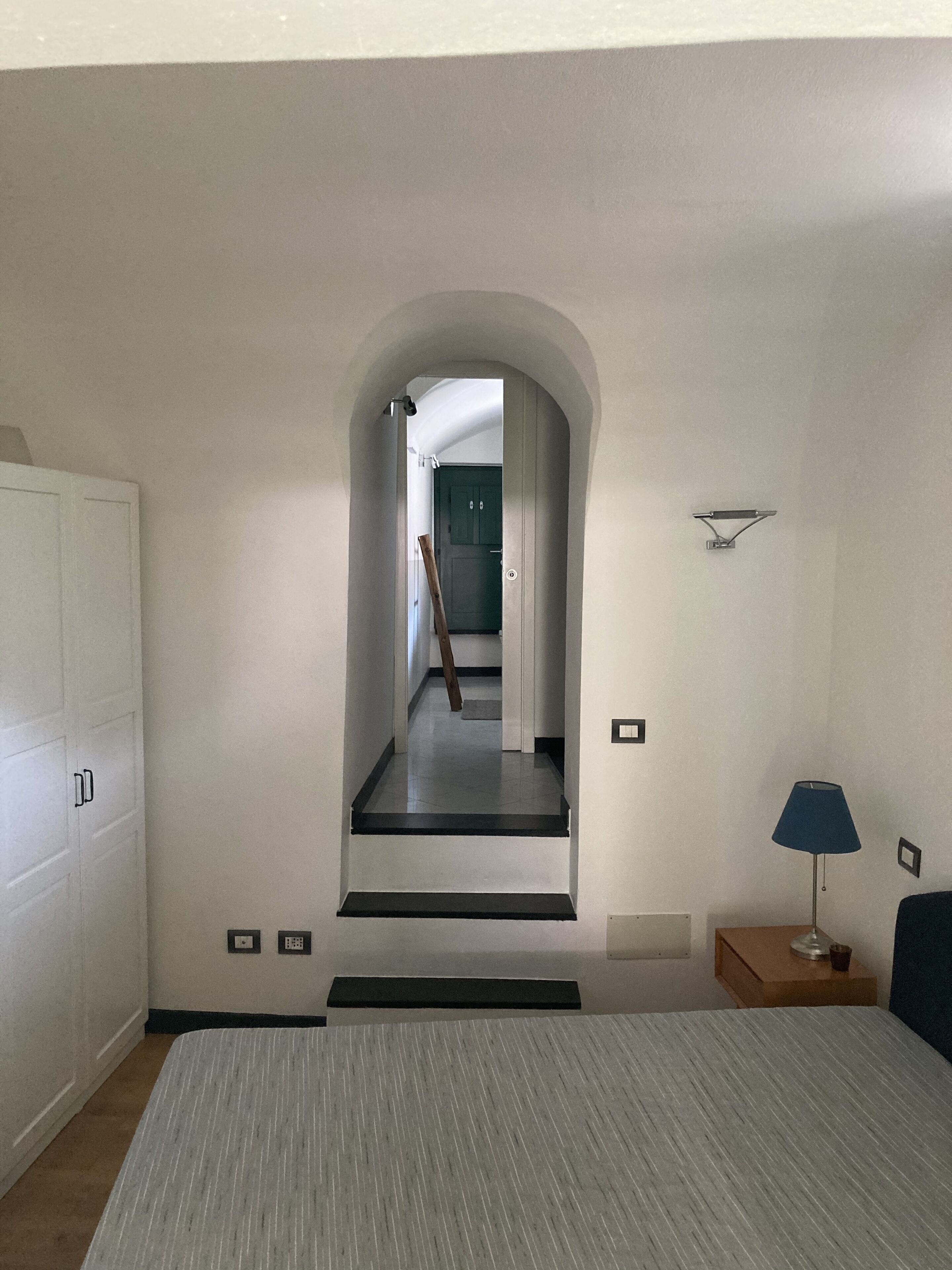Un dormitorio con cama, mesita de noche, lámpara, puerta y un arco que conduce a otra habitación.