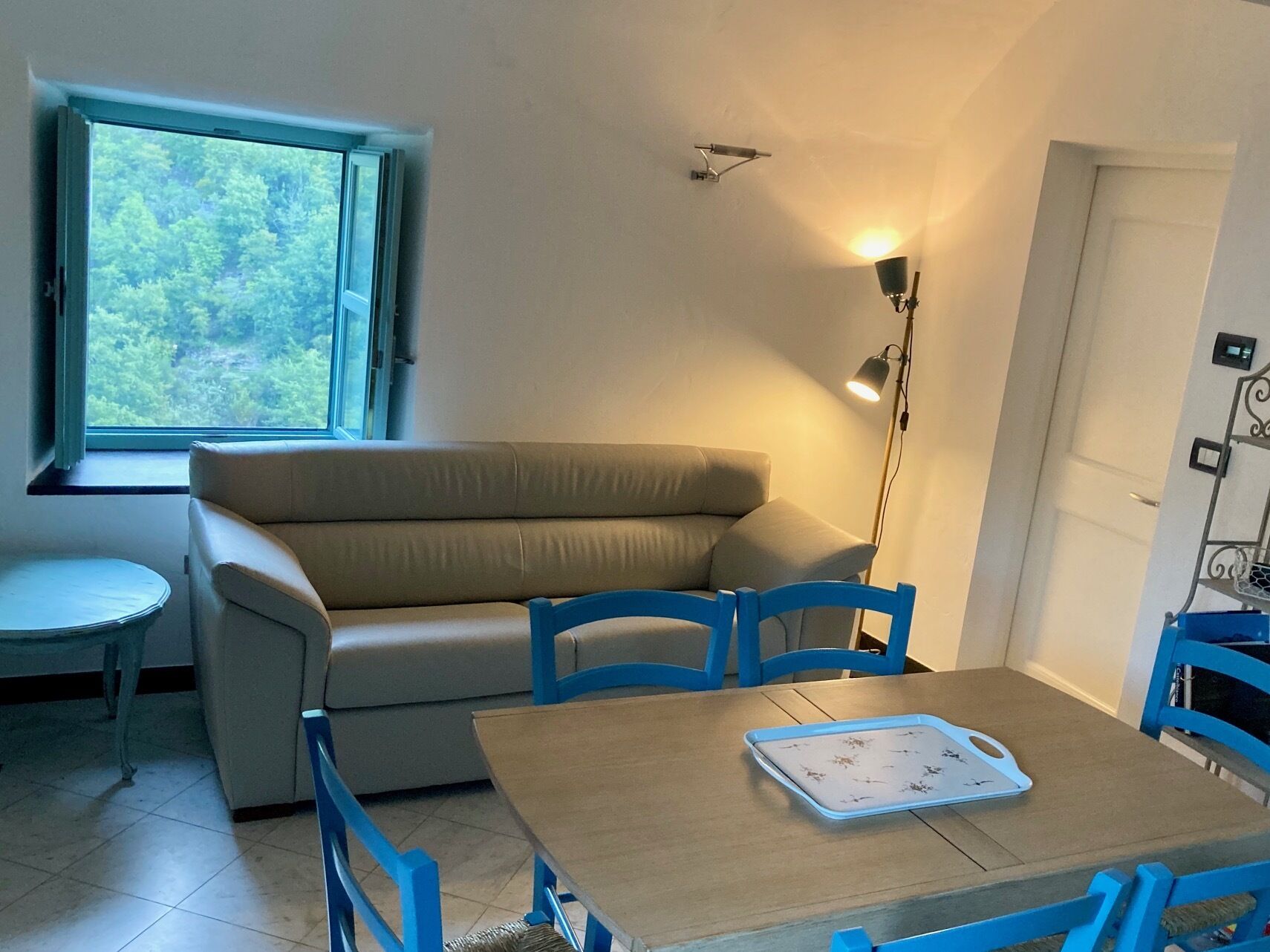Ein Zimmer mit einem beigefarbenen Sofa, zwei blauen Stühlen, einem Holztisch, einem kleinen runden Tisch, einem Fenster mit Blick auf Grünflächen und einer Stehlampe.