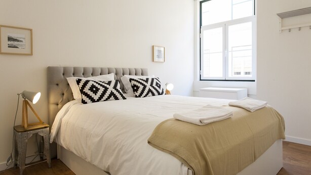 A neatly made bed with a white and beige bedspread, two patterned pillows, and a bedside lamp on a small wooden stool. - Central Downtown 3D up to 17guests by Central Hill (Lisboa ciudad)