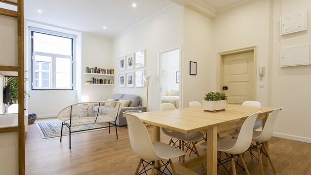 A modern living room with a wooden dining table, white chairs, a grey sofa, and a bookshelf.