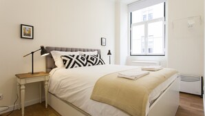 A neatly arranged bedroom with a bed, bedside lamps, a small table, and a window with a view of buildings.