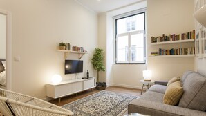 A modern living room with a sofa, a television, a bookshelf, and a large window.