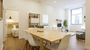 A modern living space with a wooden dining table, white chairs, a bookshelf, a TV, and a large window.