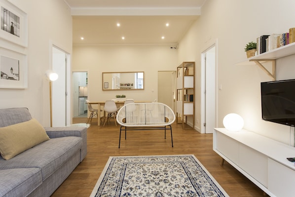 A modern living room with a sofa, a TV, a bookshelf, and a dining area in the background.
