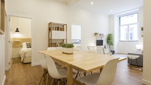 Un espacio habitable moderno con una mesa de comedor de madera, sillas blancas, una estantería, un televisor y un amplio ventanal.