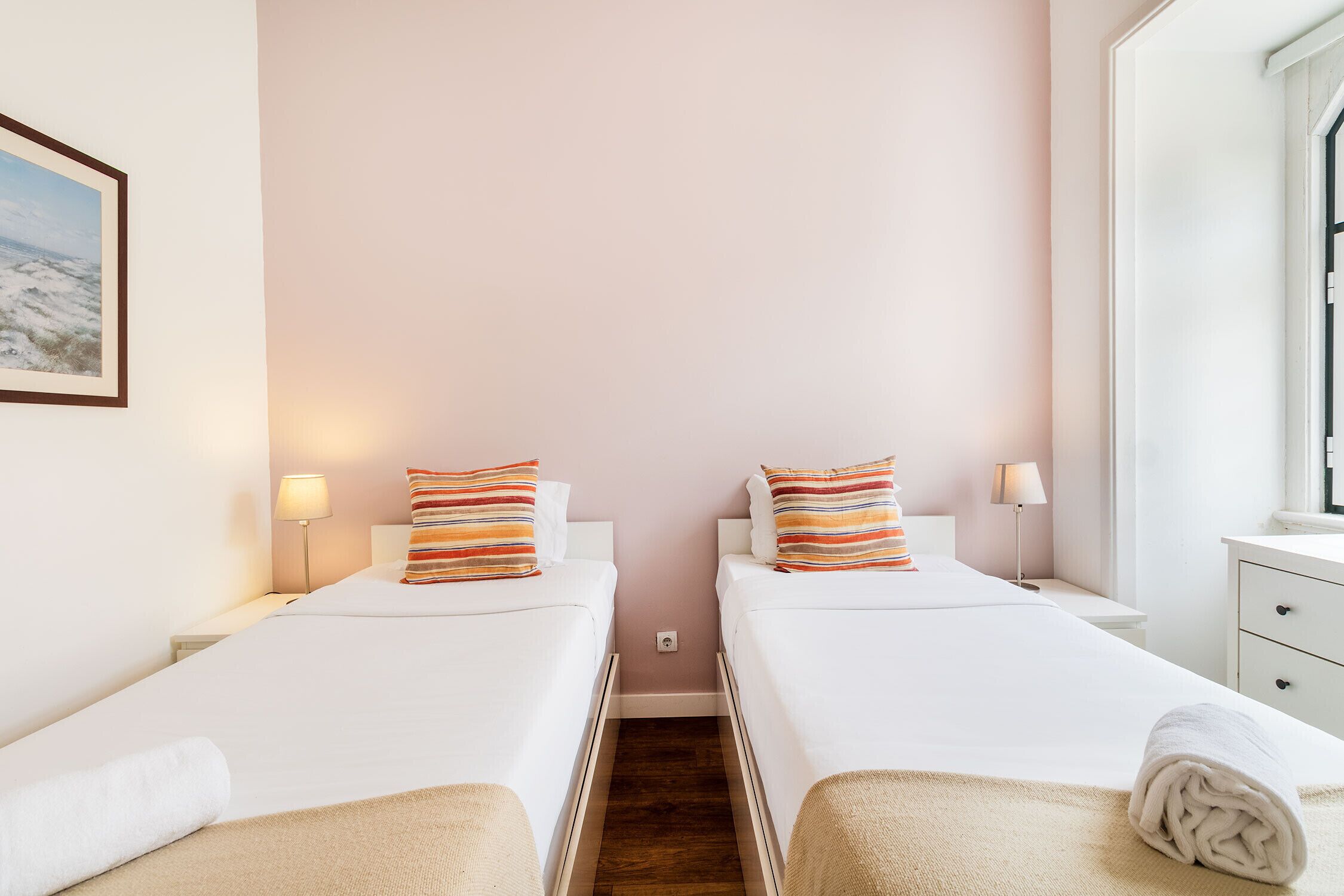 Habitación de hotel con dos camas, cada una con cubrecama blanco y almohadas a rayas, una mesita de noche con lámpara y ventana con vista a la playa.