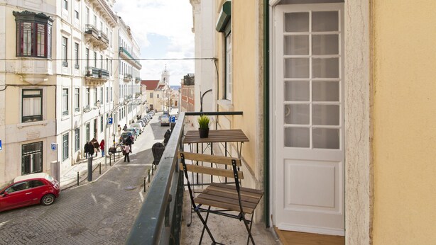 A narrow street with buildings on either side, a red car parked on the left, and a balcony with a wooden bench and a potted plant.