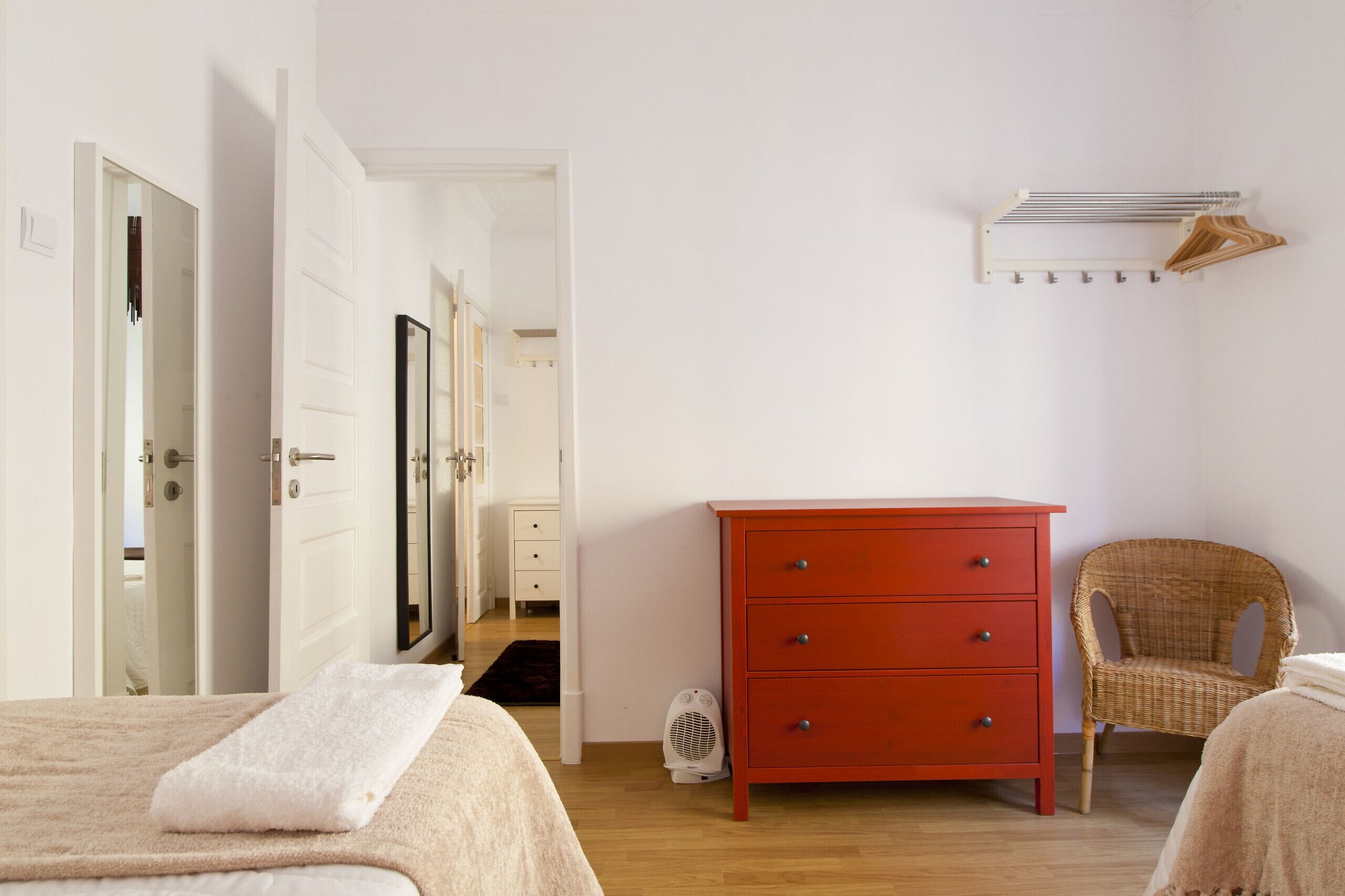 A bedroom with a bed, a red dresser, a wicker chair, and a wall-mounted drying rack.