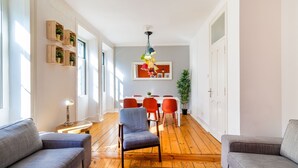 A spacious living room with a wooden floor, grey sofas, a dining area with red chairs, and a hanging light fixture.