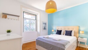 A bedroom with a large bed, wooden flooring, a window with a view, a bedside table with a lamp, and a hanging light fixture.