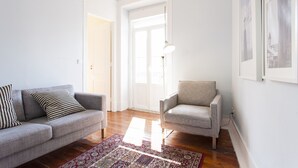 Una sala de estar con un sofá gris, un cojín estampado, un sillón gris, un tapete rojo y blanco con diseño, y un ventanal con vistas al exterior.