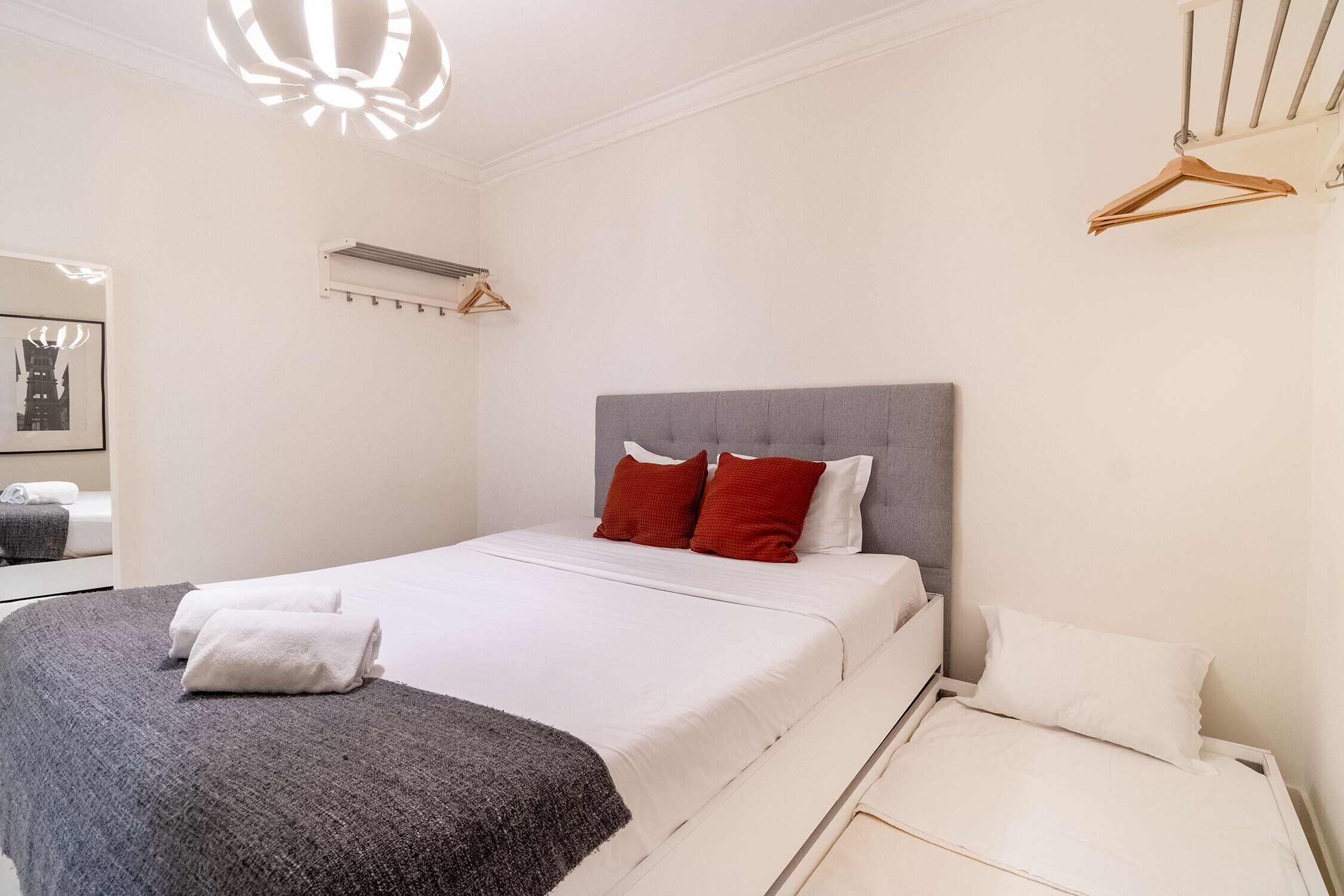 A neatly made bed with white and gray bedding, a gray headboard, and red pillows. There is a wooden hanger on the wall and a white bedside table.