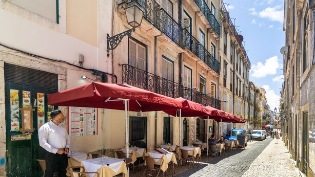 A narrow cobblestone street lined with outdoor cafes, tables, and chairs under red umbrellas.