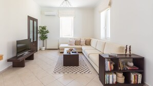 Una sala de estar moderna con un televisor de pantalla plana, una mesa de centro de madera oscura, un sofá color beige, una estantería y una planta en maceta.