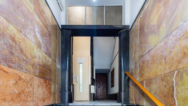 A marble-floored corridor with a wooden handrail, leading to a door with a sign.