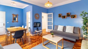 A living room with a blue wall, wooden floor, a grey sofa, a wooden coffee table, a dining table with chairs, a TV on a stand, and a plant.