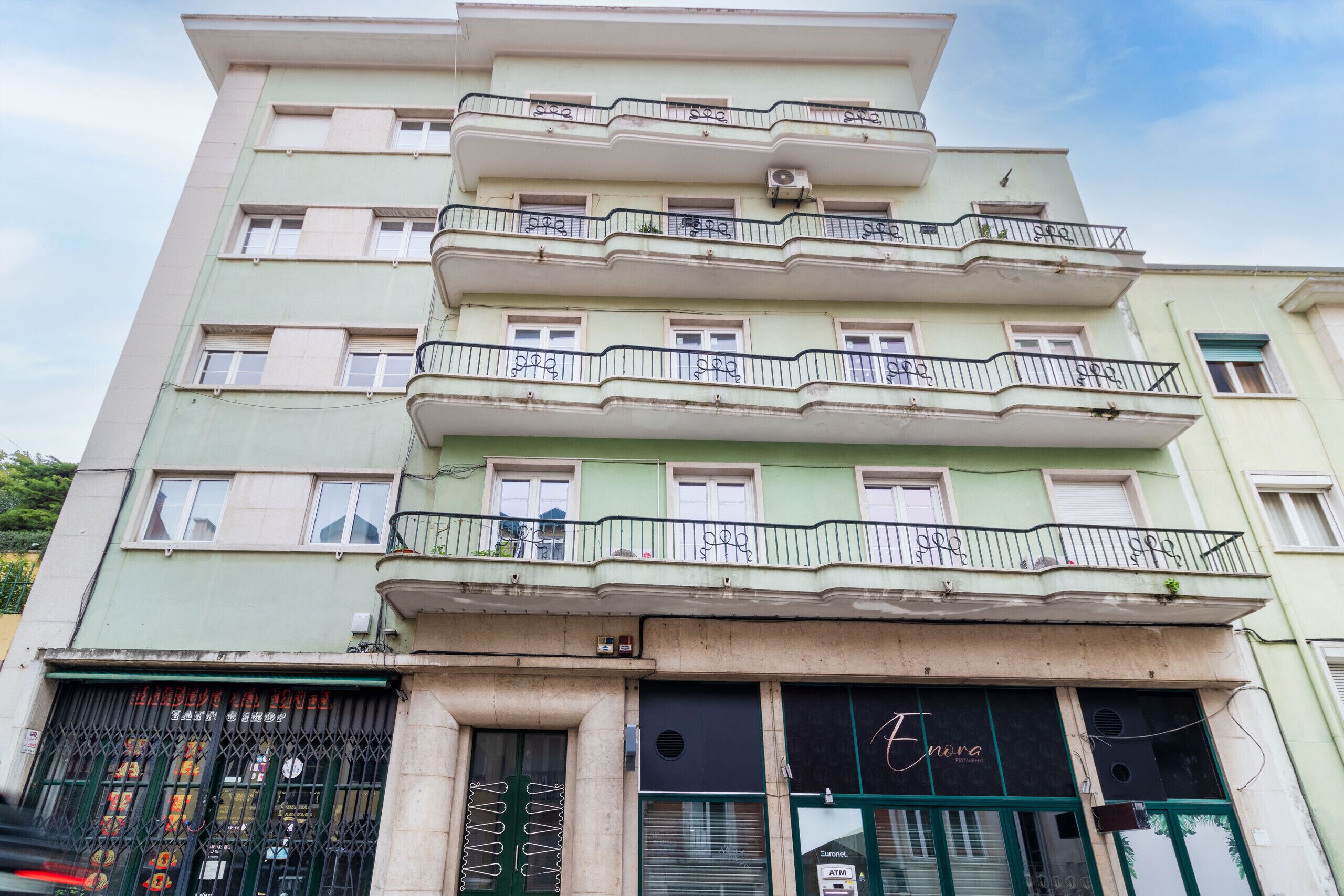 Um edifício com vários andares e varandas, restaurante chamado 'Enora' no térreo e um caixa eletrônico visível.