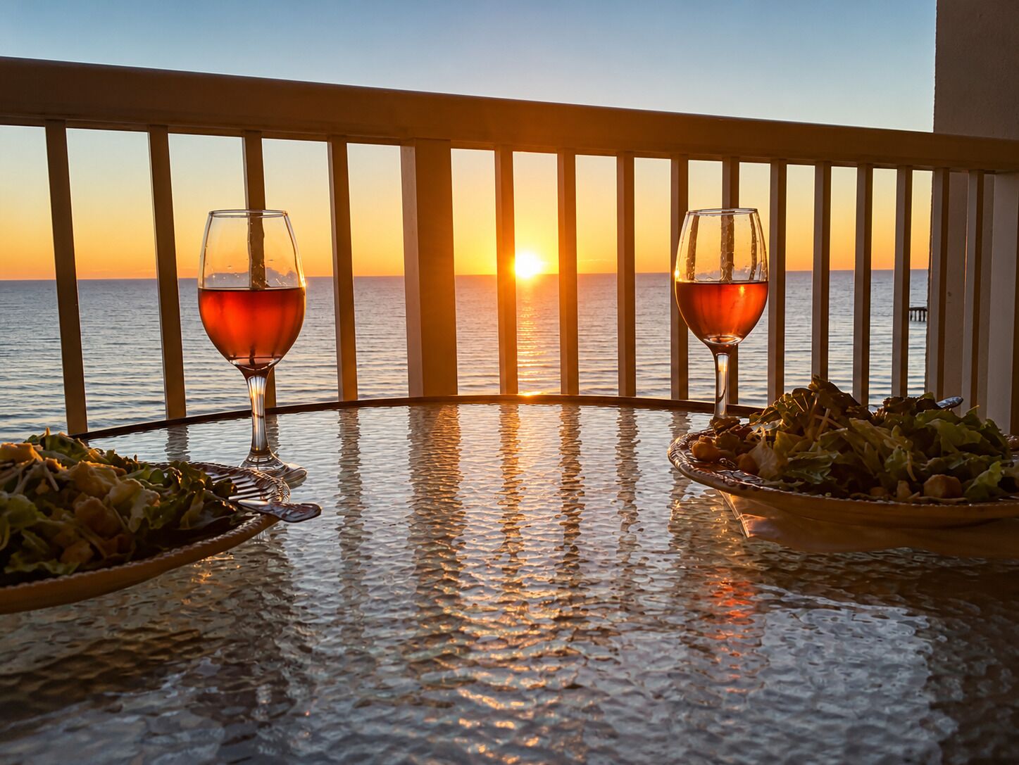 Une table avec deux verres de vin rouge et une assiette de salade, donnant sur un coucher de soleil sur la mer.