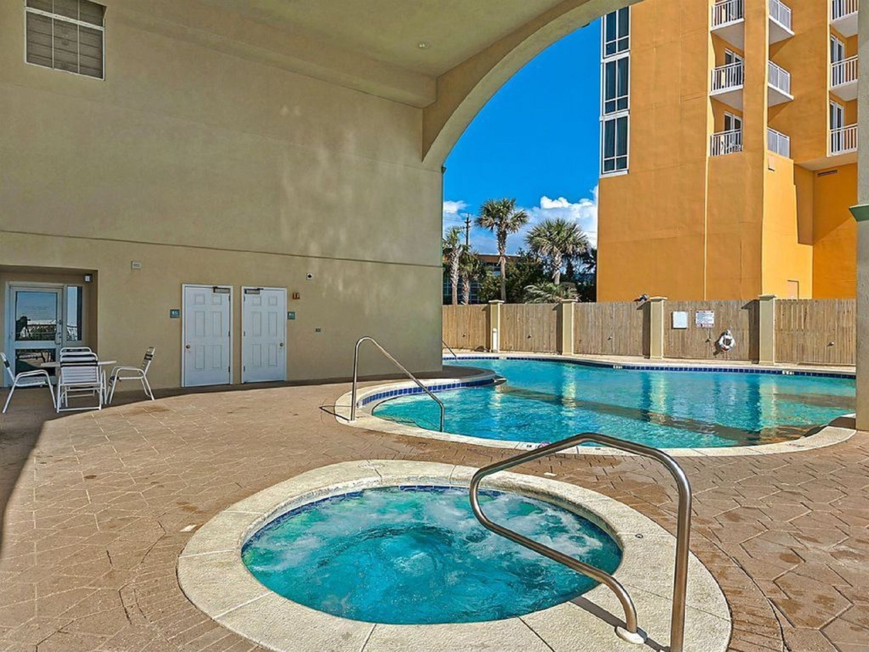 A swimming pool area with two pools, a covered seating area, and a building with balconies.