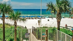 A beach with palm trees, umbrellas, and a white building. - 2 Bedroom in Ocean Villa Resort -  Beautifully decorated and appointed condo with amazing views! (Panama City Beach)